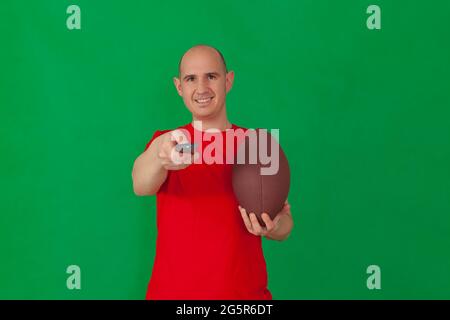 A bald caucasian man wearing a red t-shirt holds a soccer in his left hand and points a television remote control with his other hand. The background