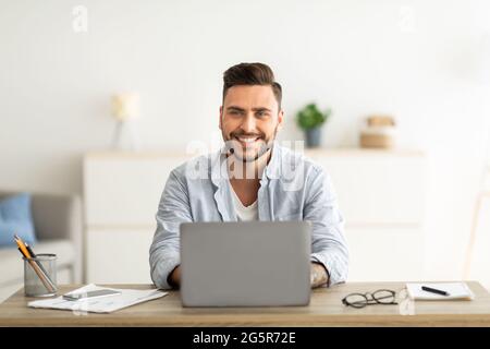 Successful freelancer. Happy man sitting at workplace with laptop computer, smiling and looking at camera Stock Photo