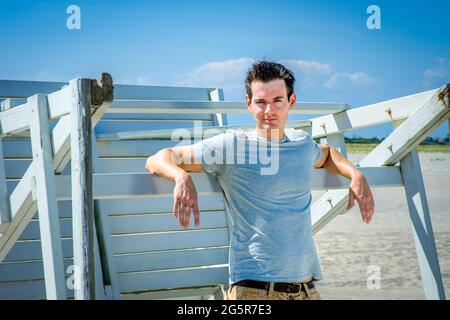 Man waiting for you. Wearing a gray t-shirt and arms resting on a wooden stick, a handsome young man is standing by a wooden structure on the beach, n Stock Photo