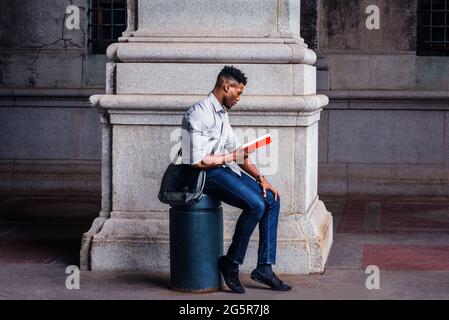 Wearing a gray shirt, blue jeans, black cloth shoes, carrying a shoulder leather bag, a young black man is sitting on a metal pillar on an old street Stock Photo