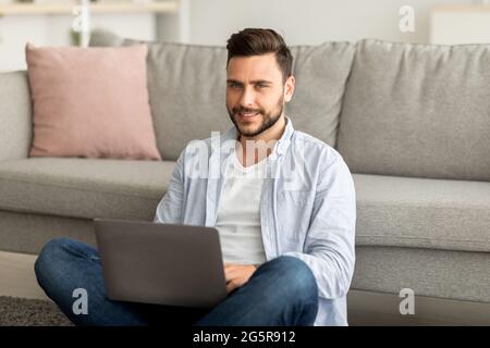 Remote job concept. Successful young freelancer man working on laptop, sitting on floor, working remotely from home Stock Photo