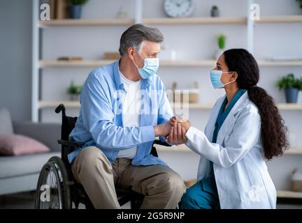 Female doctor and disabled elderly man in wheelchair wearing masks during covid lockdown, holding hands at home Stock Photo