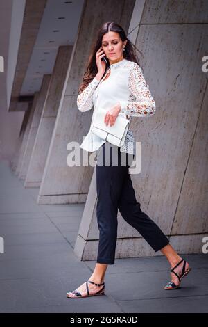 A Young Eastern European Businesswoman, wearing a white, lace cut out long sleeve shirt, black pants, sandals, holding a white leather purse, is talki Stock Photo