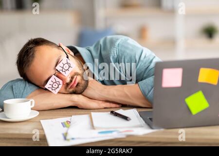 Lazy unproductive young guy wearing funny sticky notes with open eyes on his glasses, sleeping at workplace Stock Photo