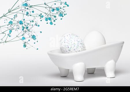 Creative experiment, composition of an egg decorated with sparkles lie in ceramic bath on white background with dry blue gypsophila flowers. Hygiene c Stock Photo