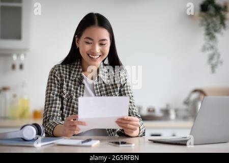 Cheerful asian woman reading letter, kitchen interior Stock Photo