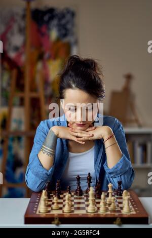 Pensive young woman thinking about chess move while sitting at home and playing chess alone Stock Photo