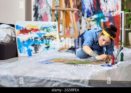 Talented female painter in apron choosing paint color, holding paintbrush while working on painting, sitting on the floor at home workshop Stock Photo