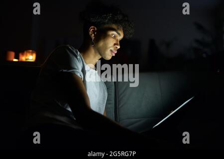 Side view of focused young guy reading e book or watching video using tablet pc at night, sitting in dark room at home Stock Photo