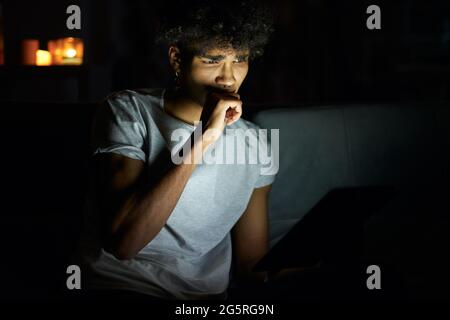Portrait of young guy looking concentrated while reading e book using tablet pc at night, sitting in dark room at home Stock Photo