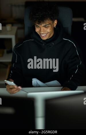 Technology. Stylish young guy with piercing smiling while using smartphone, sitting at the table and working on laptop late in the evening Stock Photo
