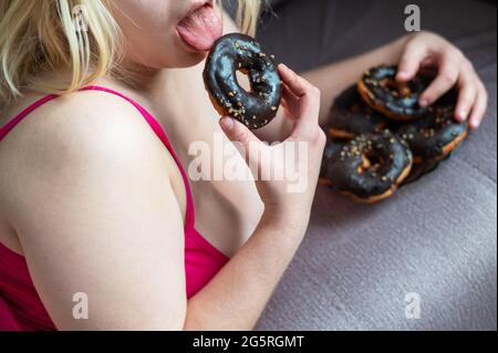 Fat blonde eats a chocolate donut. Unrecognizable obese young woman enjoying sweet buns for breakfast. Cropped photo. Stock Photo