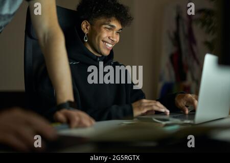 Love my job. Cheerful young guy with piercing smiling while sitting in front of laptop screen and working late in the evening Stock Photo