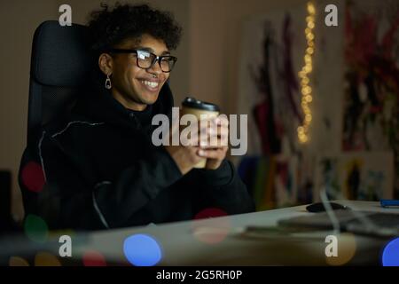 Leisure activities. Happy young guy with piercing wearing glasses holding disposable cup of coffee, sitting at the table and watching videos using Stock Photo