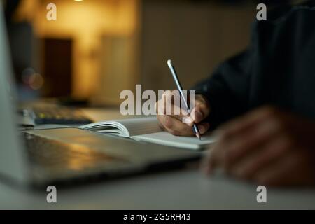 Writing ideas. Close up shot of hands of male student making notes in his notebook while studying, using laptop late at night Stock Photo