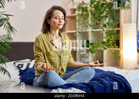 Relaxed young woman sitting on bed in Lotus pose, meditating at home Stock Photo