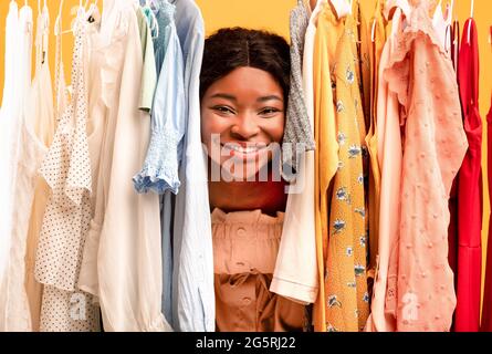 Young black woman peeking out between clothes hanging in rail, smiling at camera, satisfied with shopping choice Stock Photo