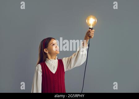 Younger schoolgirl looks at the light bulb, which she raised up standing on a gray background. Stock Photo