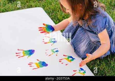 Little girl 2-4 draws leaving rainbow colors child's handprints on large white sheet of paper Stock Photo