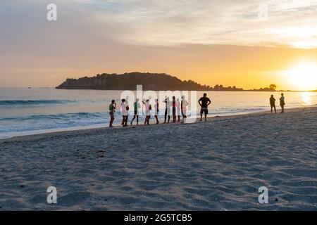 Mount Maunganui New Zealand -January 20 2015; Group of young people standing at waters edge waiting to enter as golden glow of rising sun warms beach. Stock Photo