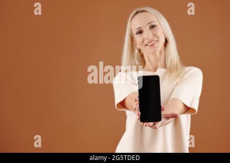 Positive young blond-haired woman in tshirt giving new smartphone with blank screen to you, studio shot Stock Photo
