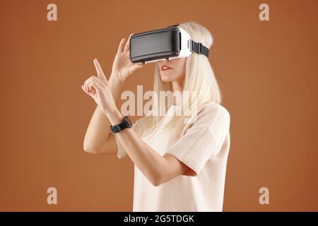 Modern curious young blond-haired woman in tshirt standing against brown background and touching invisible board while using virtual reality device Stock Photo