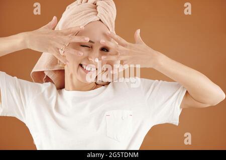 Portrait of smiling playful young Caucasian woman with hair wrapped into towel keeping fingers spread against face while using beauty product Stock Photo