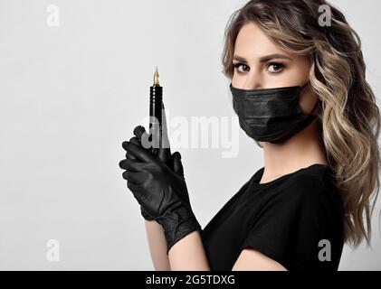 Blonde curly woman in black protective medical mask and latex gloves stands sideways holding permanent makeup machine Stock Photo