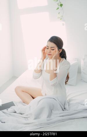 Miserable young woman touching temples sitting on bed at home, Stock Photo