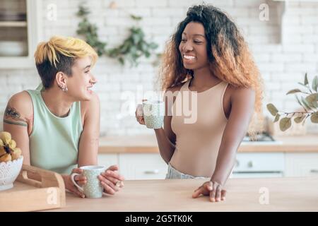 Two cheerful girlfriends talking in kitchen with coffee Stock Photo