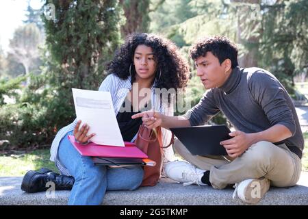 Two latin students studying together sitting on a bench outdoors. University life at campus. Stock Photo