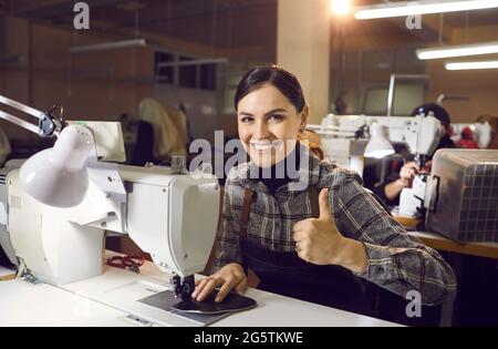 Satisfied factory worker sitting at shoe sewing machine, giving thumbs up and smiling Stock Photo
