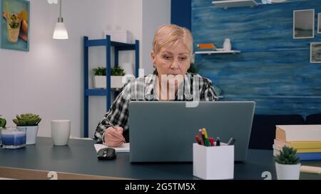 Serious mature middle aged business woman using laptop typing email and writing on notebook, working at home office, focused senior old lady searching information on internet or communicating online Stock Photo