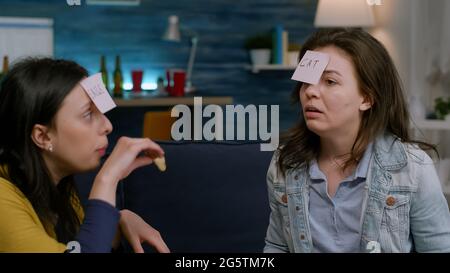 Group of multiethnic people playing interesting guess who game putting sticky notes on forehead eating snacks having fun during evening party. Multiracial friends hanging out together late at night Stock Photo