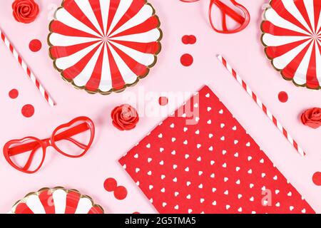 Party flat lay with paper plates, heart shaped glasses, roses, confetti and drinking straws on pink background Stock Photo