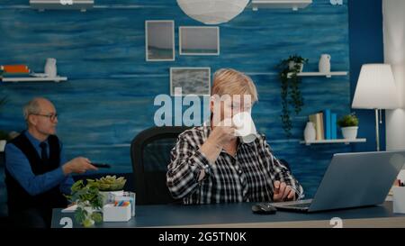 Older professional mature business woman using laptop sitting at workplace desk drinking coffee. Retired remote executive manager working from home typing on pc, while husband watching tv in background Stock Photo