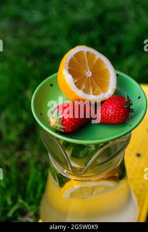 Defocus saucer with lemon and two strawberry standing on glass jug of lemonade with slice lemon and leaves of mint on yellow board. Blurred grass Stock Photo