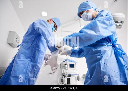 Side view of assistant in blue surgical suit helping surgeon to put on sterile gloves. Two doctors in protective medical masks preparing for plastic surgery in clinic. Concept of medicine and surgery. Stock Photo