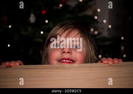 Little girl peeks over a table with lit christmas tree in the background Stock Photo