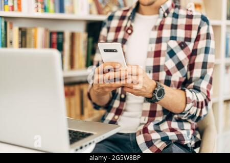 Close-up of a man with a laptop, uses a mobile phone, types a text message, a student is studying while sitting in the library against the background Stock Photo
