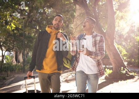 Two happy mixed race male friends carrying luggage walking in city park talking and using smartphone Stock Photo