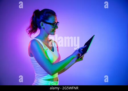 Holding tablet, crazy excited. Caucasian young woman's portrait on black studio background in neon light. Beautiful male model in white shirt. Stock Photo