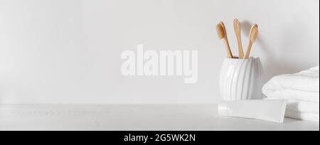 Minimal bath banner with bamboo toothbrushs in ceramic glass, white tube of toothpaste and white towels on white background. Front view. Copy space Stock Photo