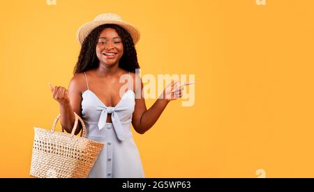 Charming black woman in dress and hat holding straw bag, pointing aside at free space over orange background, banner Stock Photo