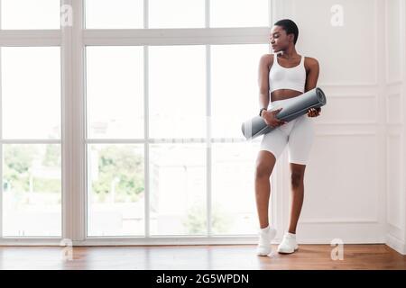 Young black woman holding yoga mat standing near window Stock Photo