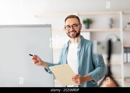Young male teacher conducting online lesson near blank blackboard, explaining new material to students, copy space Stock Photo
