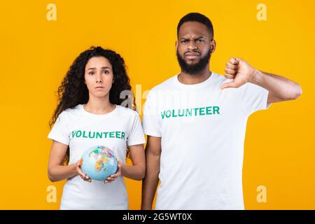 Female And Male Volunteers Holding Globe Gesturing Thumbs-Down, Yellow Background Stock Photo