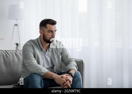 Thoughtful serious bearded millennial male sit on sofa alone at home Stock Photo