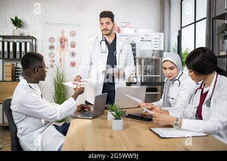 Two men and two women in white lab coats with stethoscopes sitting at table and using modern gadgets. Multiracials doctors analyzing together diagnosis of severe patient. Stock Photo