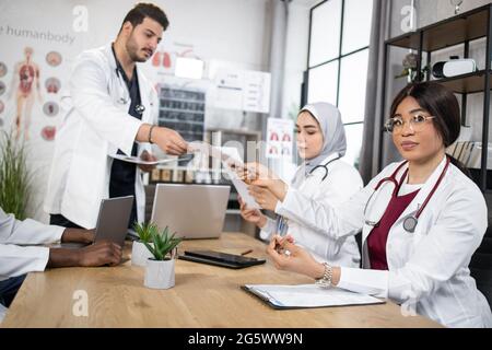 Two men and two women in white lab coats with stethoscopes sitting at table and using modern gadgets. Multiracials doctors analyzing together diagnosis of severe patient. Stock Photo
